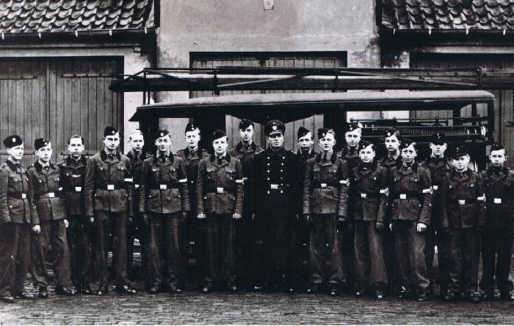 Feuerwehr – Hitlerjugend Telgte, Jahrgänge 1925 bis 1928 aufgenommen am 03.01.1943 Im Hintergrund ist das Spritzenhaus von 1898 zu sehen, welches 1960 abgerissen wurde, und der erste Mannschafts- und Gerätewagen der Feuerwehr Telgte, der 1931 in Dienst gestellt wurde.